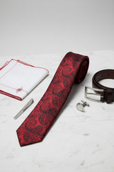 Red paisley silk tie displayed with cufflinks and pocket square on marble surface.